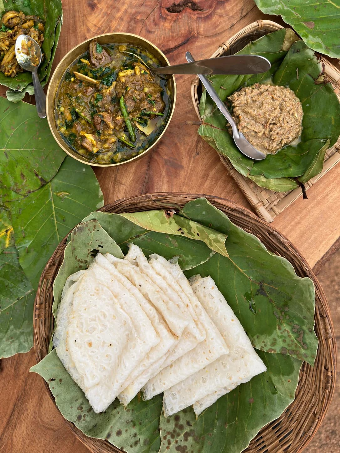 Folded white food on leaves in a woven basket, with accompanying dishes on a wooden table at Ajam Emba